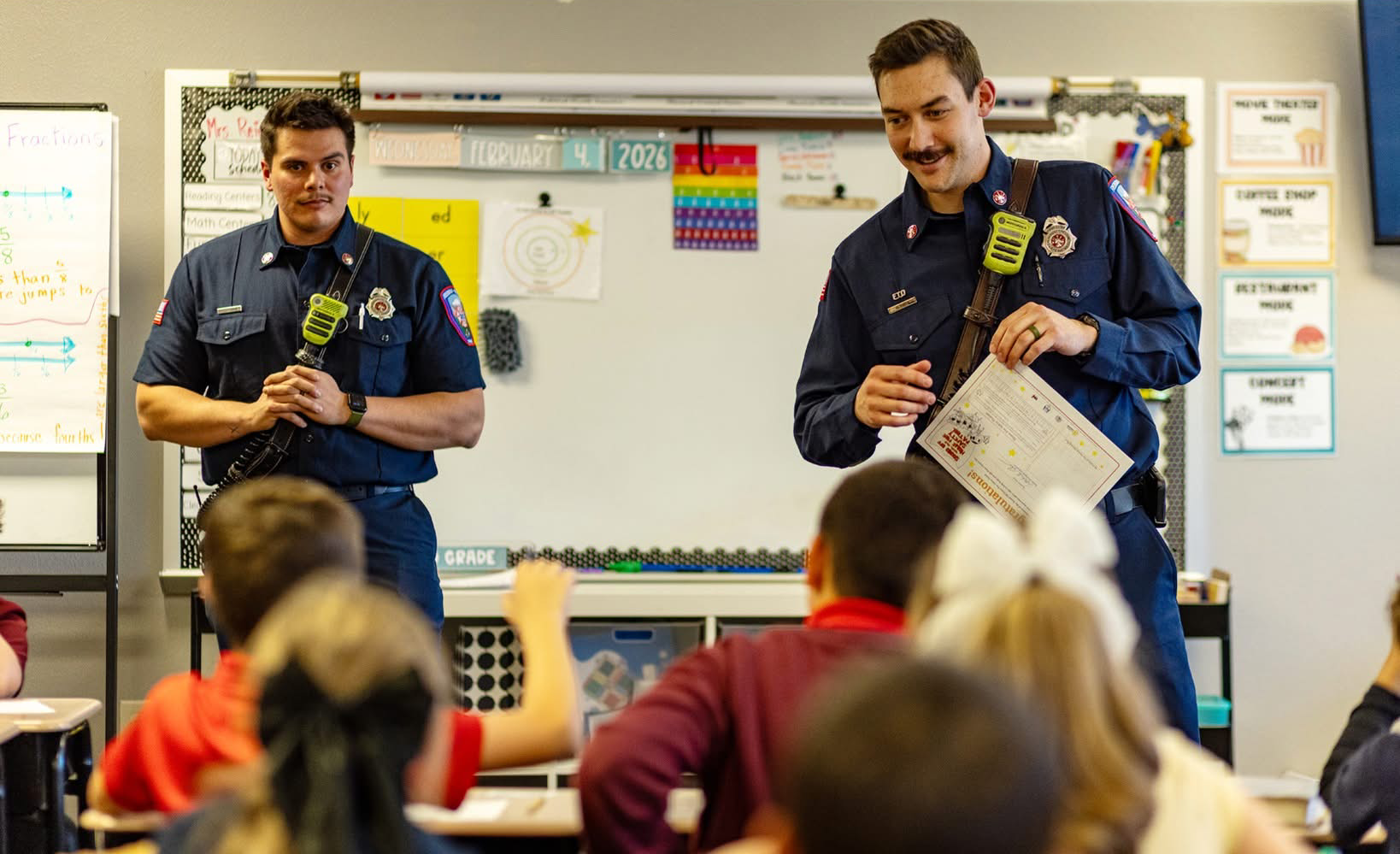 Two firefighters are engaging with students in a classroom, sharing information and possibly teaching a lesson.