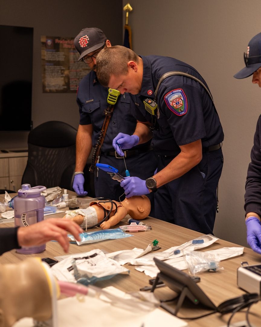 Emergency responders practice medical procedures on a training mannequin at a table filled with supplies and equipment.
