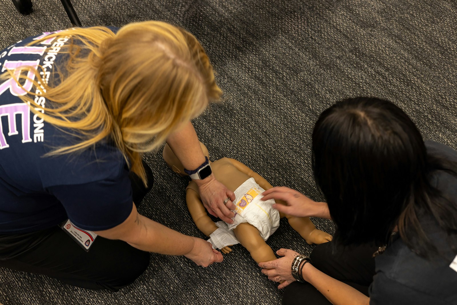 Two people are practicing how to change a diaper on a baby doll during a training session.
