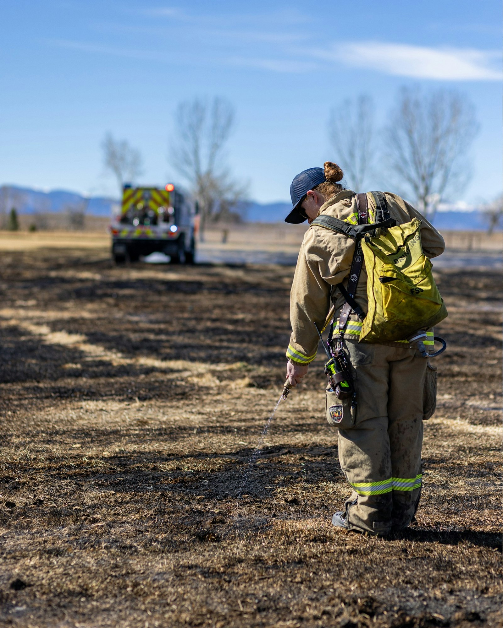A firefighter in protective gear sprays water on a burnt area, with a fire truck visible in the background on a clear day.