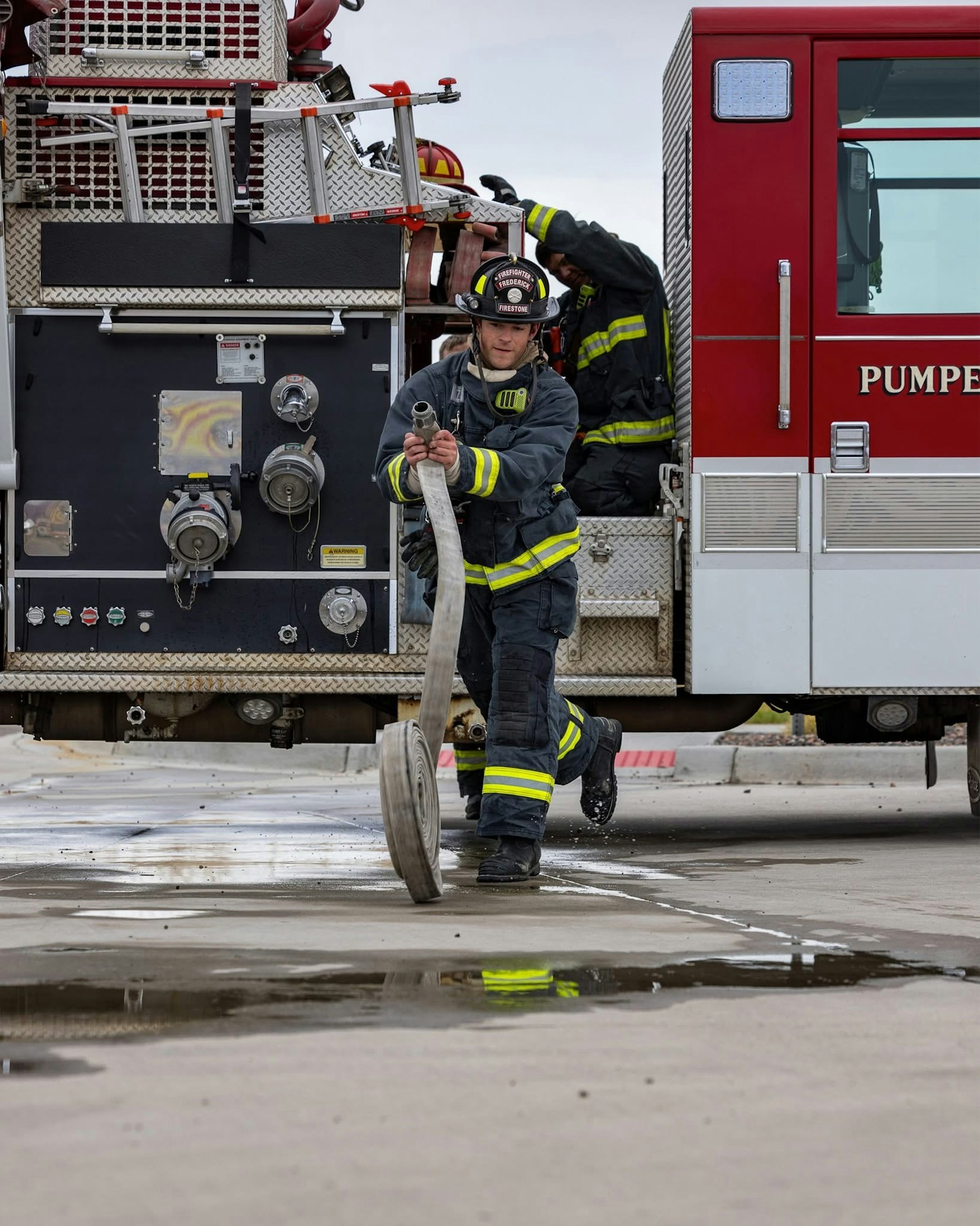 A firefighter is pulling a fire hose from a fire truck, while another firefighter is visible inside the vehicle.