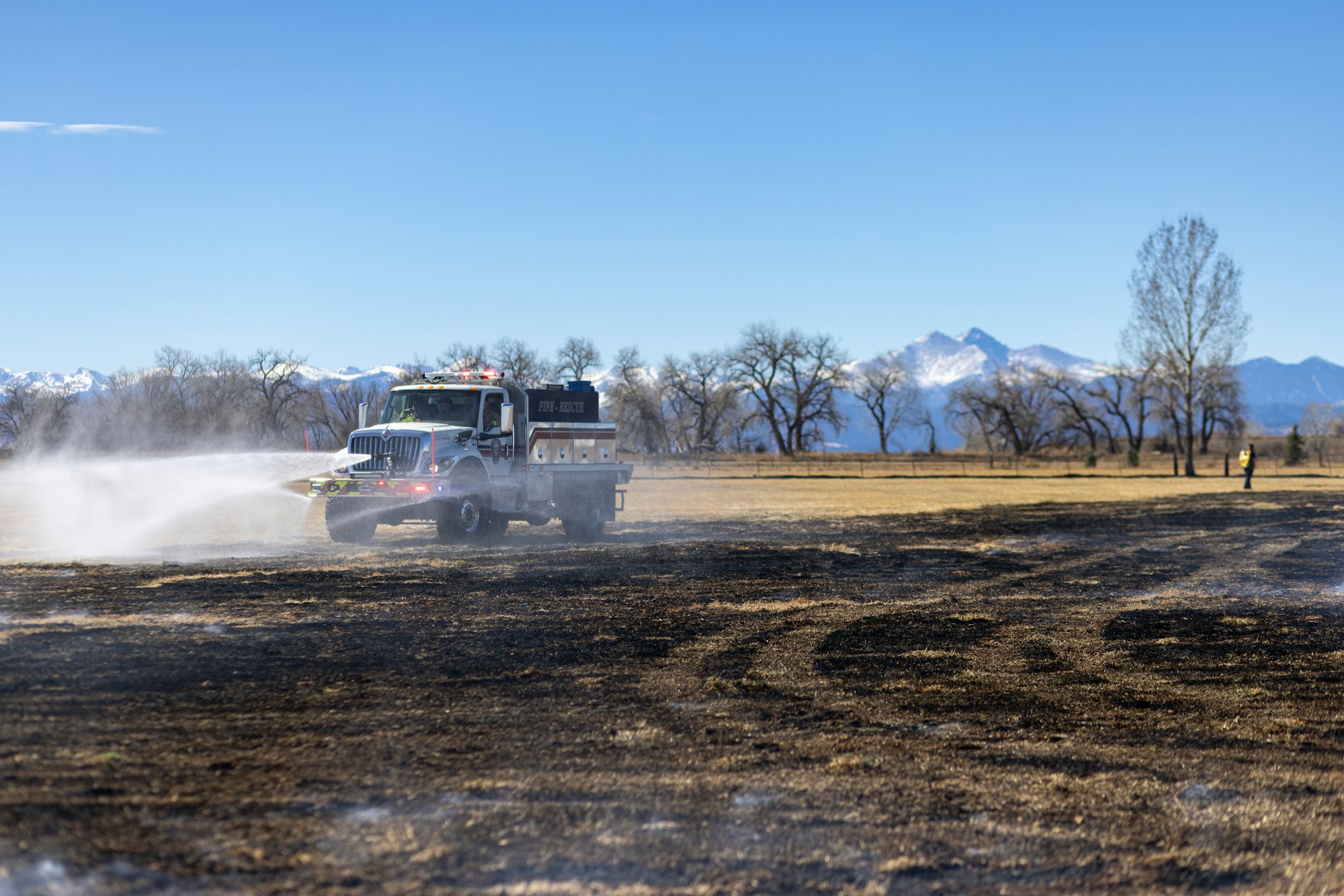 A fire truck sprays water on a burned field, with mountains in the background and a person standing nearby.