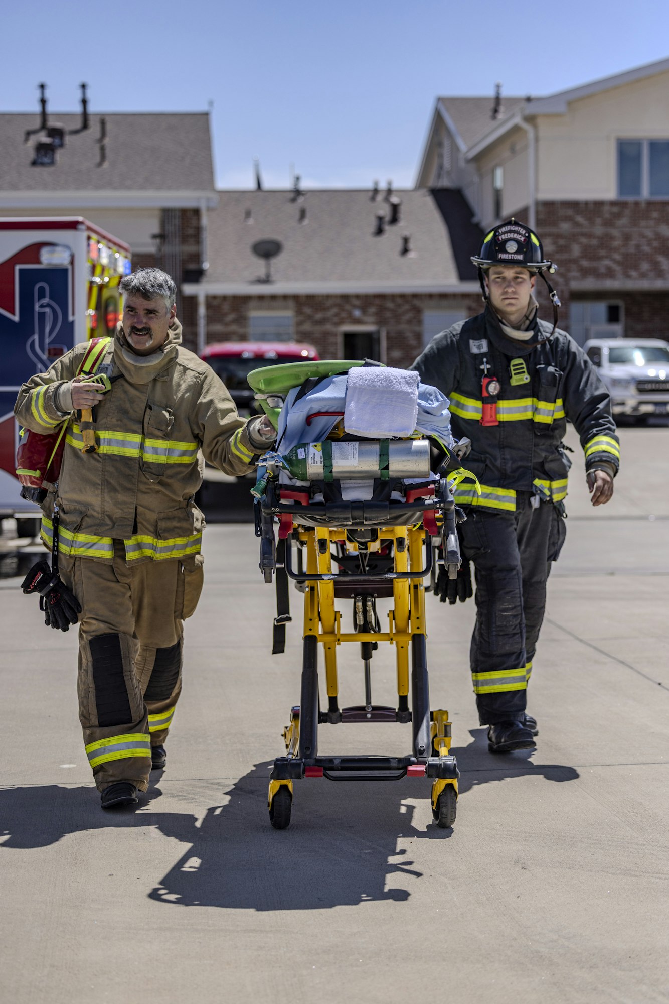 Two firefighters are transporting a stretcher with medical equipment in a residential area, near an ambulance.