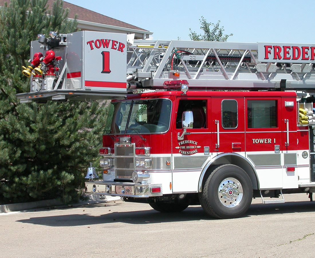 A red firetruck with "TOWER 1" labeled on its side, parked beside trees.
