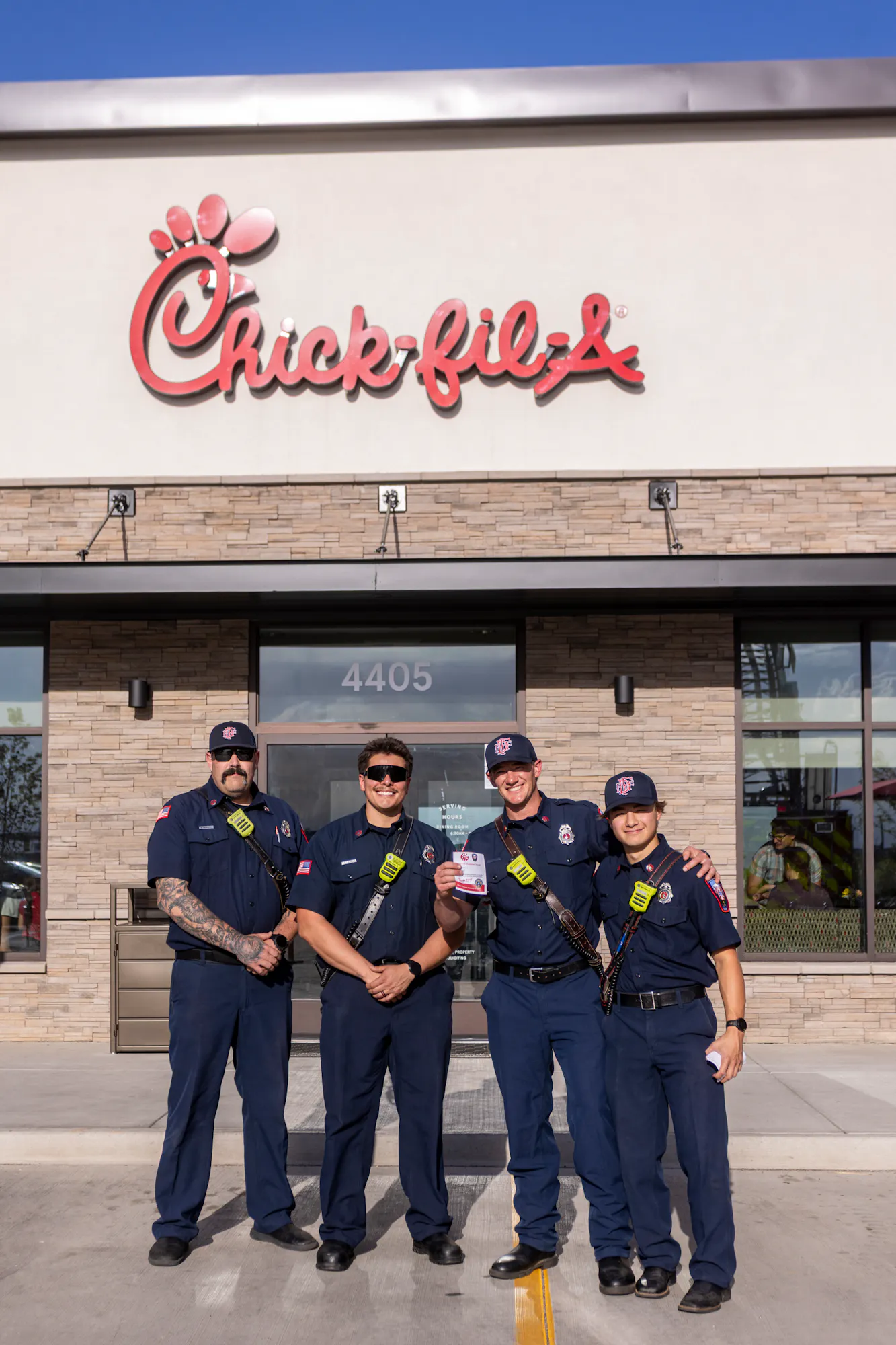Four firefighters posing outside a Chick-fil-A restaurant, smiling and holding something, with the restaurant's sign visible.