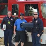 Three people in front of a fire truck, one holding a case, smiling.