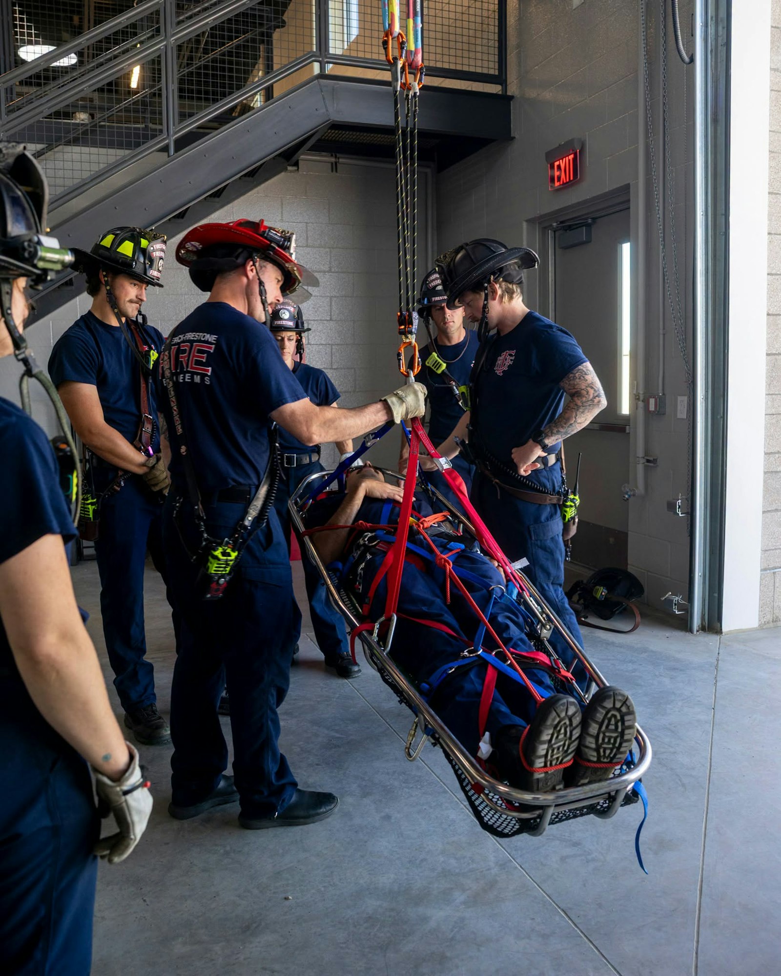 Firefighters are training to rescue a person using a stretcher and ropes in a facility. Safety gear and teamwork evident.