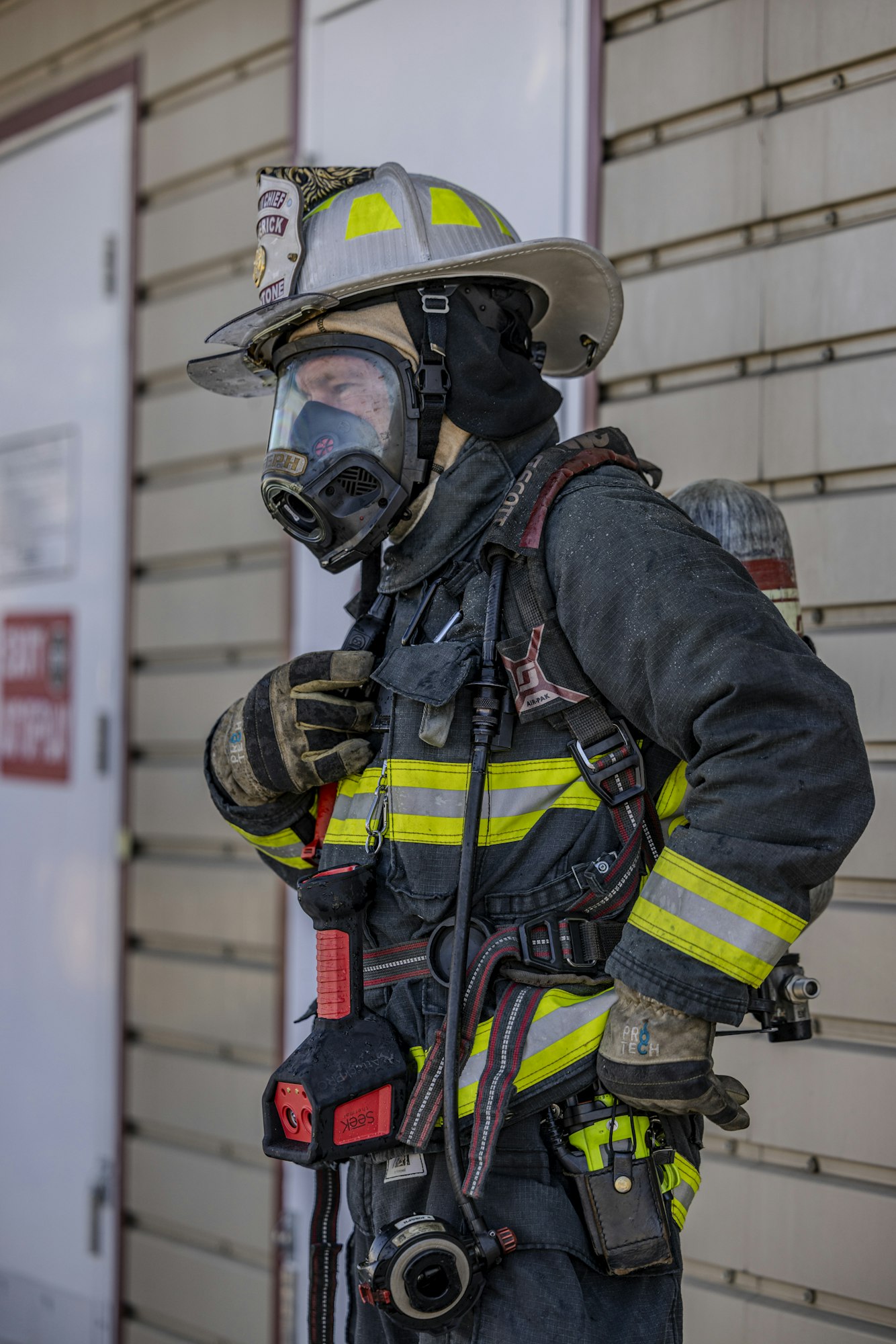 A firefighter in full gear, including a helmet, mask, and harness, stands near a building, prepared for action.