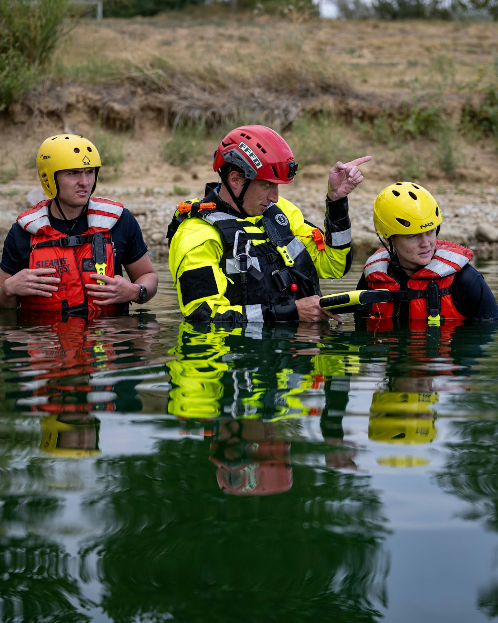 Three individuals in safety gear are standing in calm water, engaged in what appears to be a discussion, possibly about rescue operations.