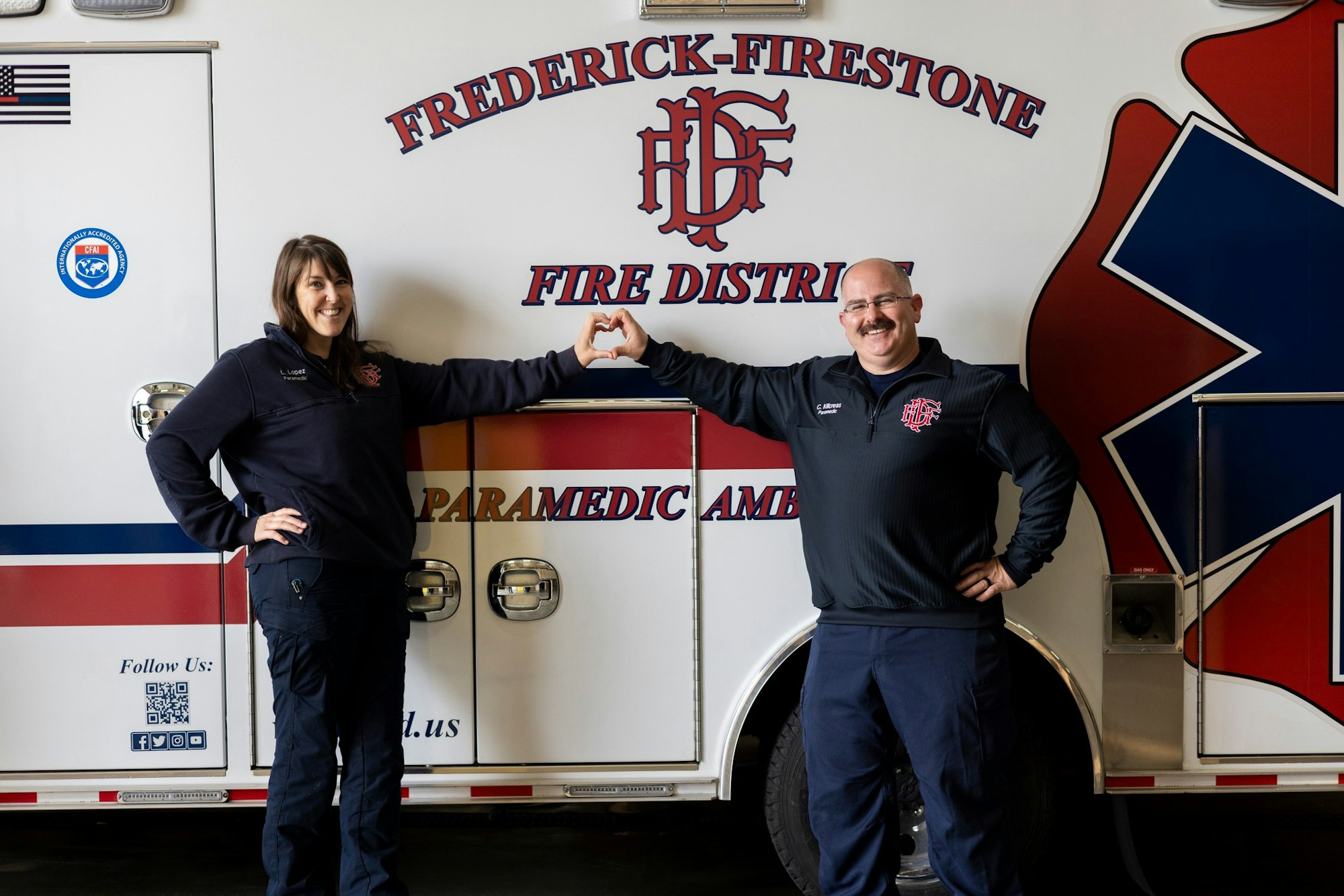 A man and woman in firefighting gear are making a heart shape with their hands in front of a fire department ambulance.