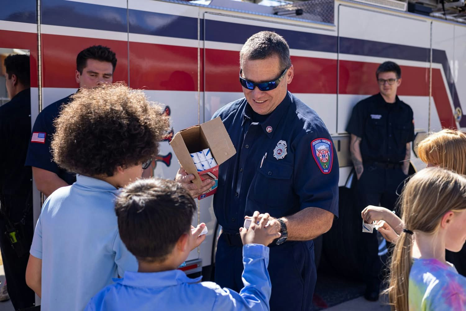 A firefighter hands out items to children, with others in uniforms and smiles, in front of a fire truck. Community engagement event.