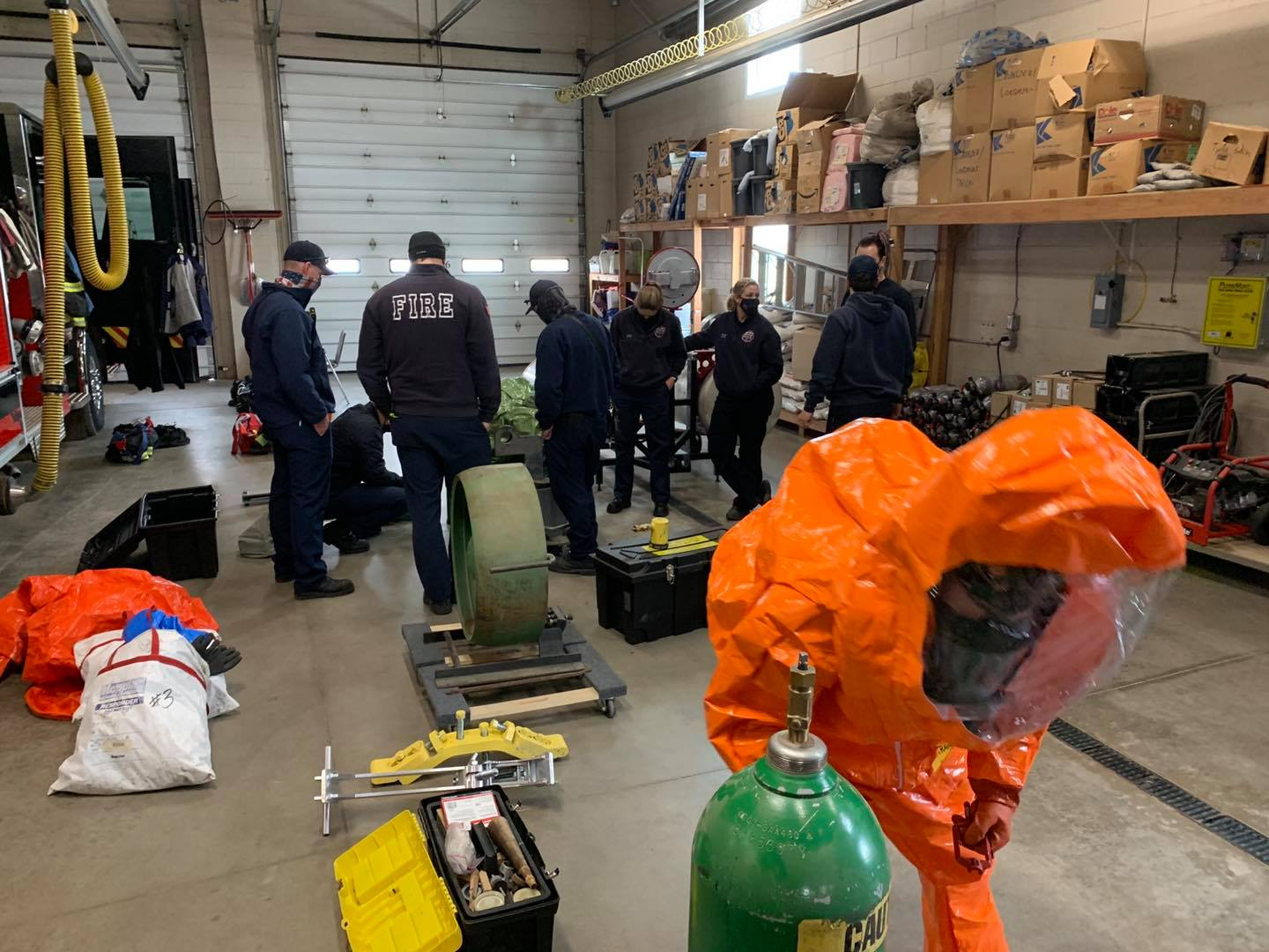 Firefighters in a garage; one in hazmat gear. Equipment scattered around, possibly a training scenario.