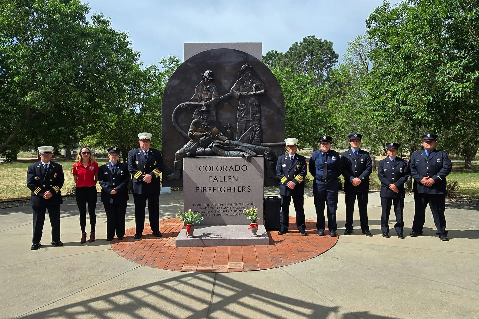 A group of firefighters and a woman stand by a memorial honoring Colorado's fallen firefighters, surrounded by greenery.