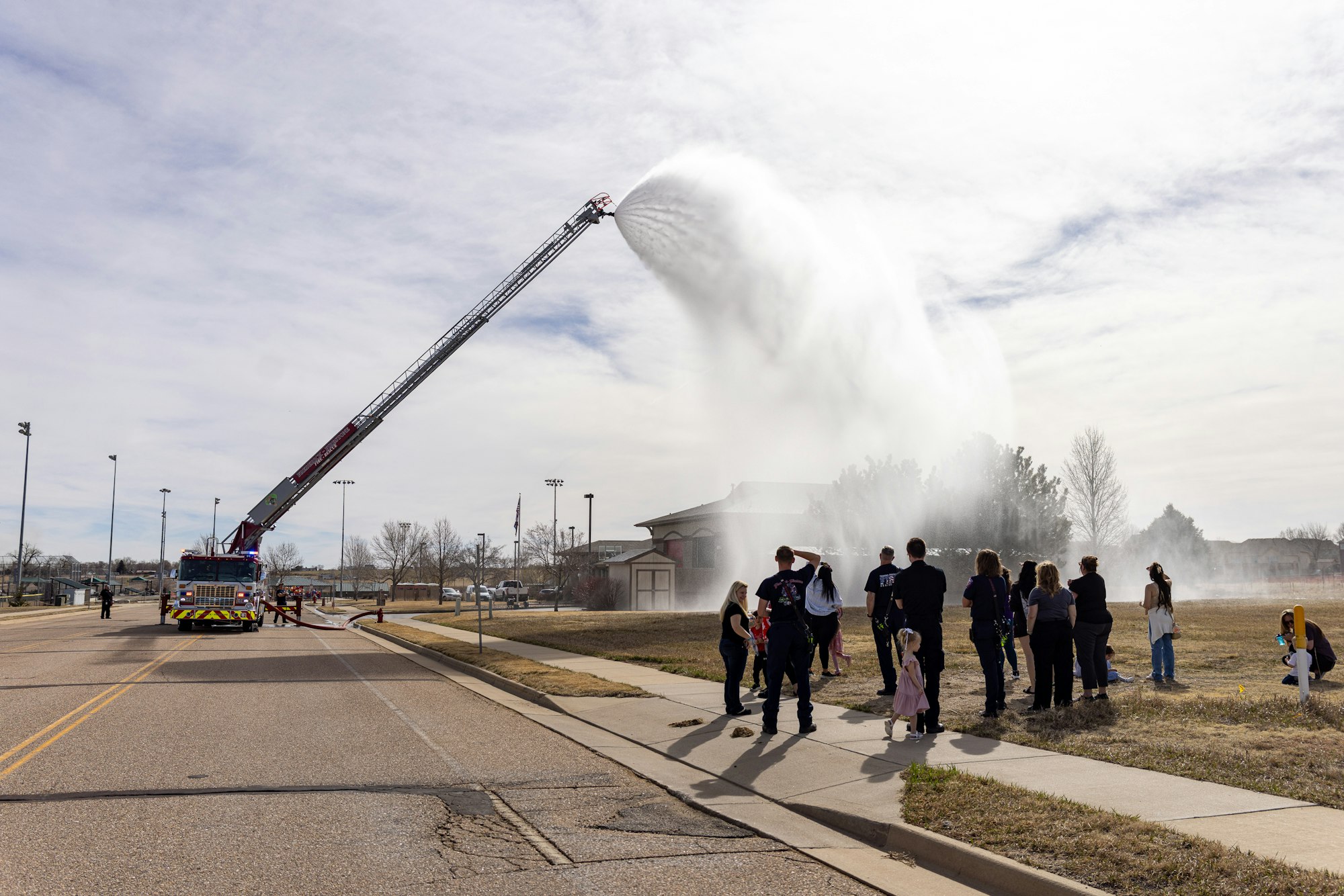 A fire truck sprays water high into the air while a crowd watches nearby, against a clear sky.
