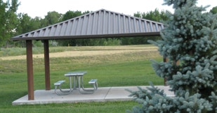 A park shelter with a picnic table, surrounded by greenery.