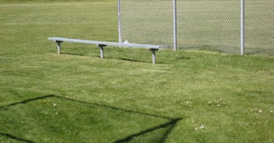 A metal bench on grass by a chain-link fence, with shadows cast on the ground.