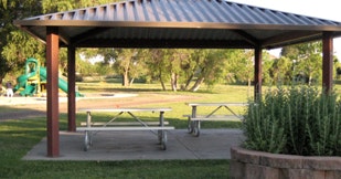 A park shelter with a picnic table, greenery, and a playground in the background.