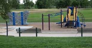 A playground with a climbing wall, swings, and other equipment, surrounded by grass and a fence.