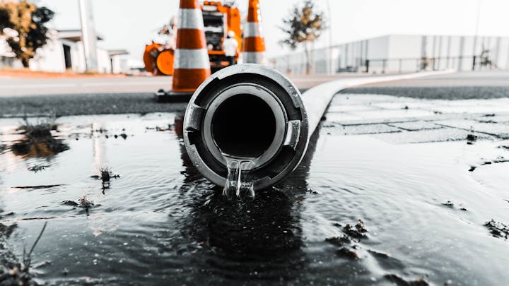A large hose lying on the wet pavement, leaking water from its opening. In the background, there are orange traffic cones and construction equipment, suggesting a work zone or emergency response area.