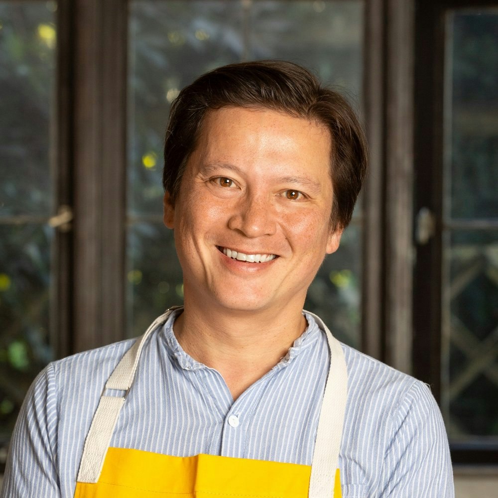 A person smiling, wearing a striped shirt and a yellow apron, standing indoors.
