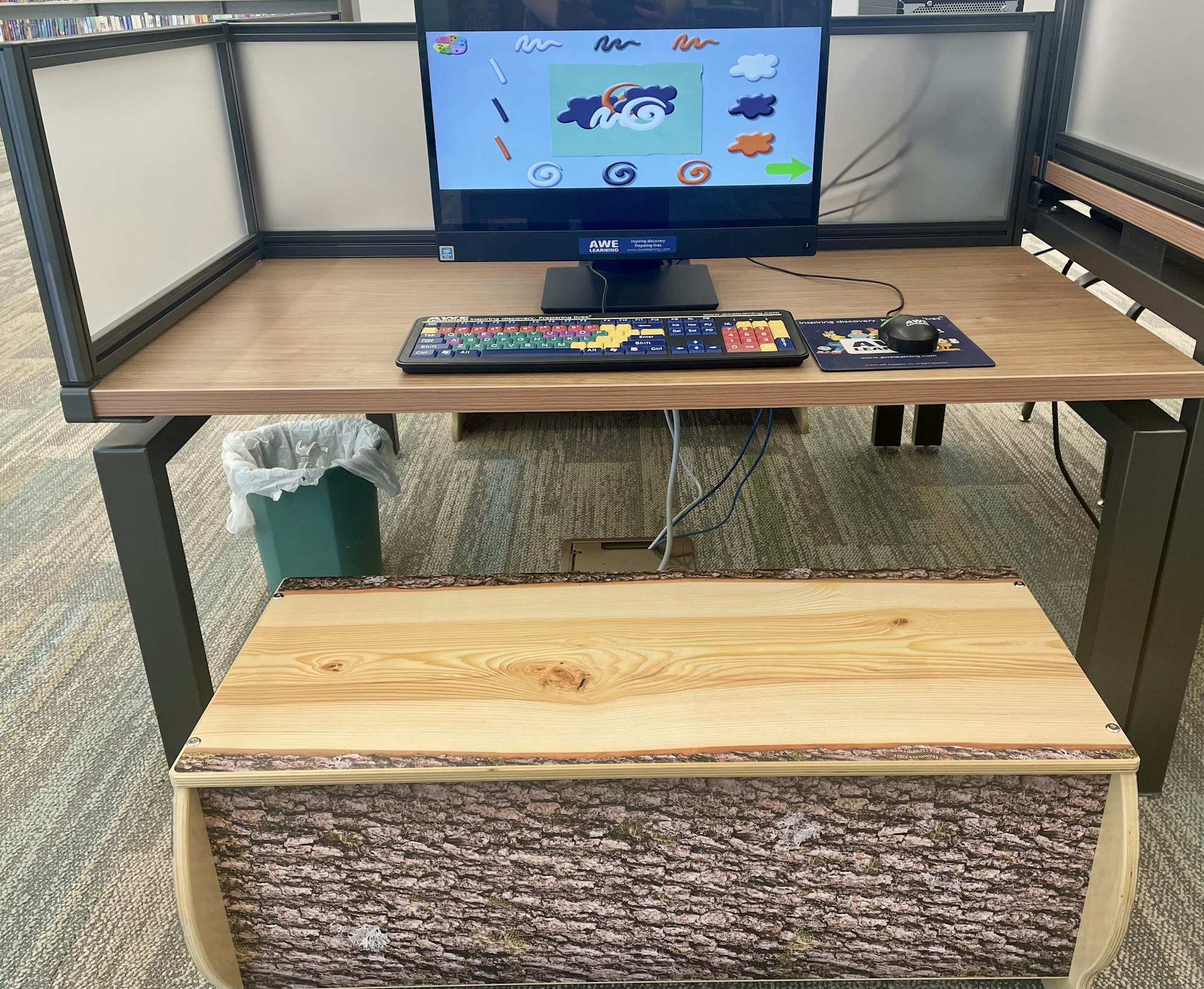 Computer station with a colorful keyboard and kid-friendly educational program on screen; wood-themed bench and trash can nearby.