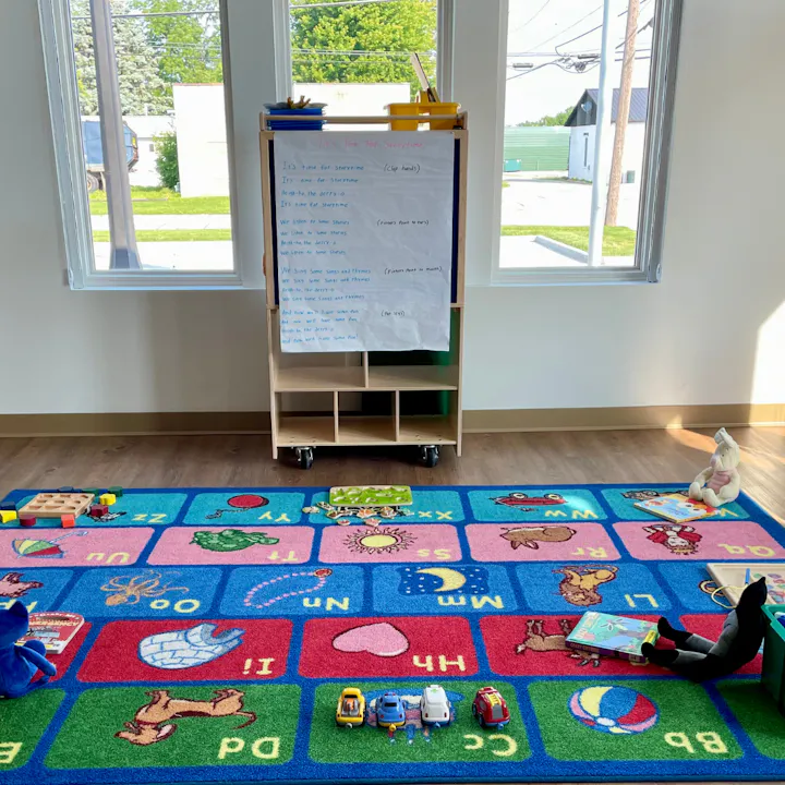 A classroom with a colorful alphabet rug, toys, books, and a whiteboard easel with writing.