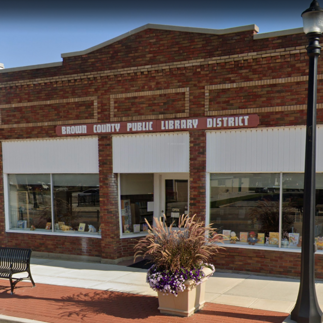 A brick library building with large windows, an "Open" sign, a bench, and a planter with flowers outside.