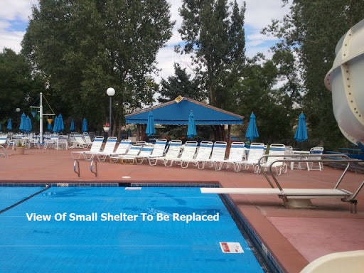 A swimming pool area featuring lounge chairs, blue umbrellas, and a small shelter slated for replacement.