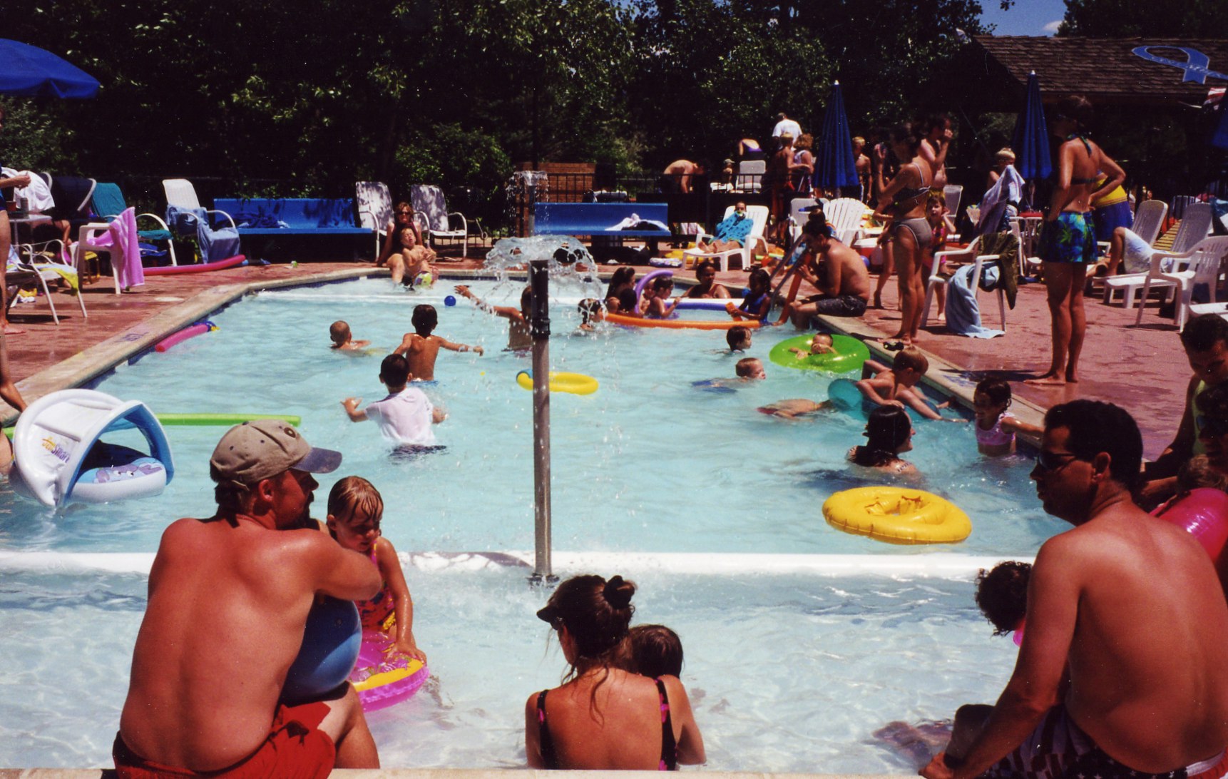 A busy pool scene with children playing, adults relaxing, and floats scattered in the water on a sunny day.