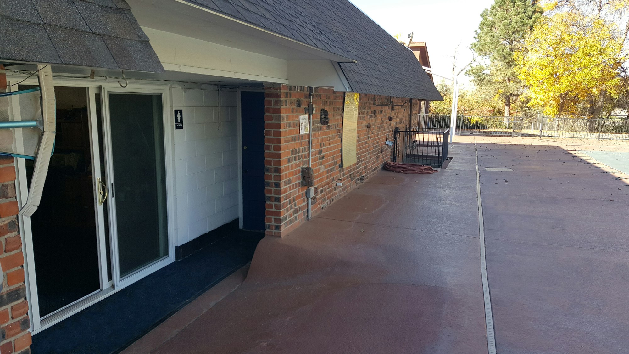 The image shows a brick building exterior with a patio area, a sliding door, and a sign for a women’s restroom.