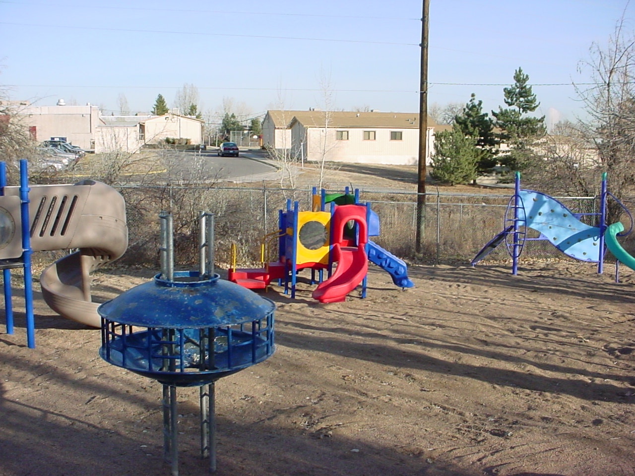 A playground featuring colorful slides, climbing structures, and equipment, surrounded by dirt and some trees.