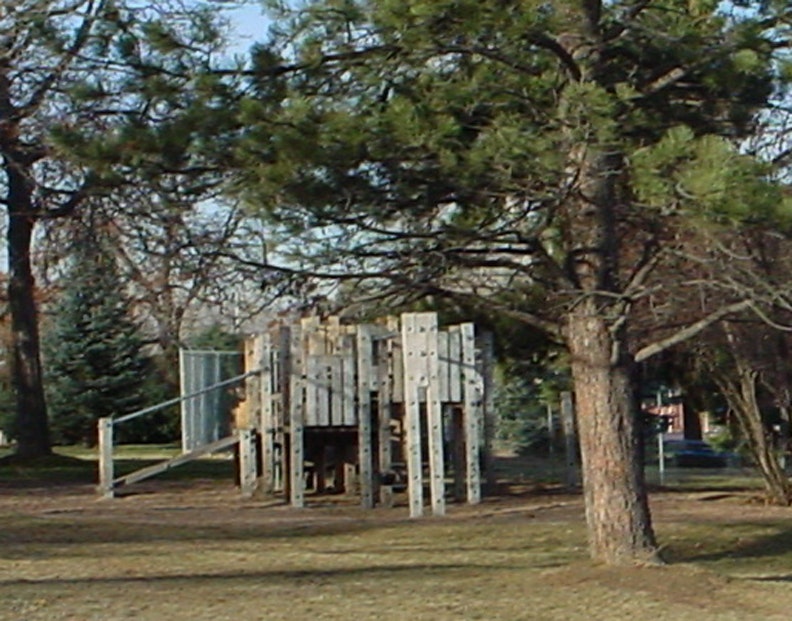 A wooden playground structure surrounded by trees and grassy area in a park setting.