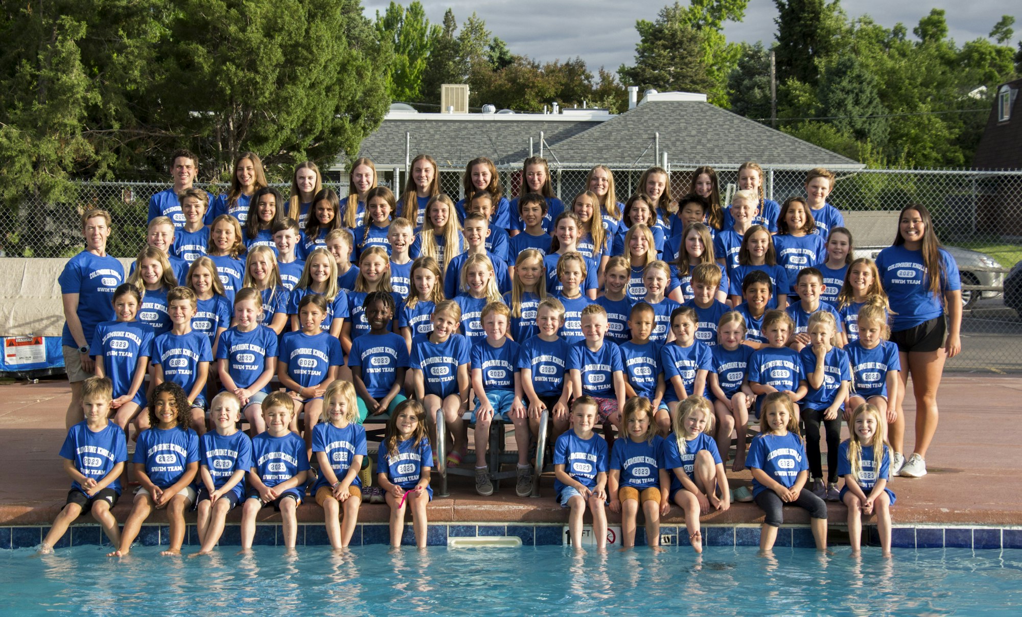 Group of children and adults in blue t-shirts labeled "Swim Team" by a pool.