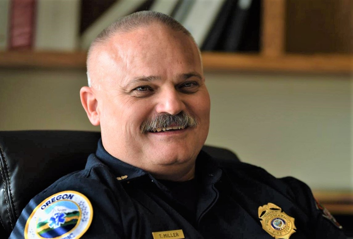 Fire Chief Tom Miller pictured in his navy blue uniform, seated in his office, smiling.
