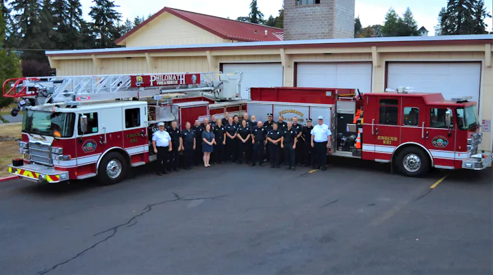 A group of firefighters poses in front of fire trucks at a fire station, showcasing their vehicles and camaraderie.