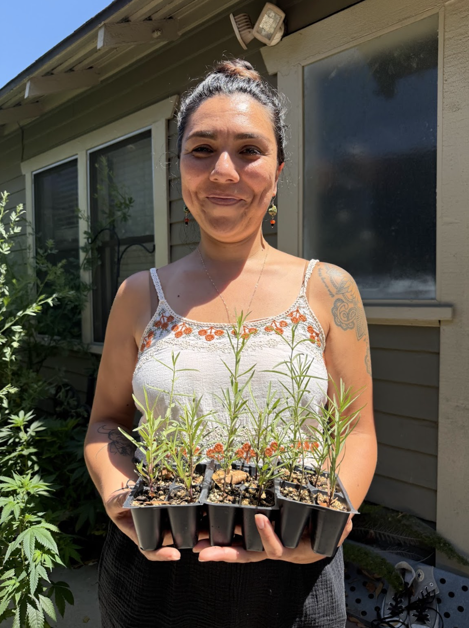 A woman is holding a tray of young plants outside, smiling in bright sunlight, surrounded by greenery.