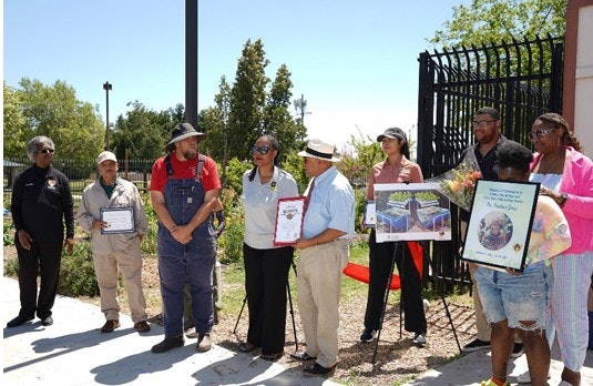 A group of people stands outdoors, holding certificates and photos, likely at a community event or ceremony.