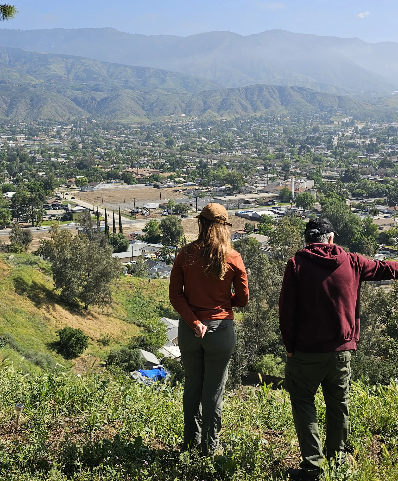 Two people stand on a hillside overlooking a scenic view of a town, with mountains in the background and greenery around.