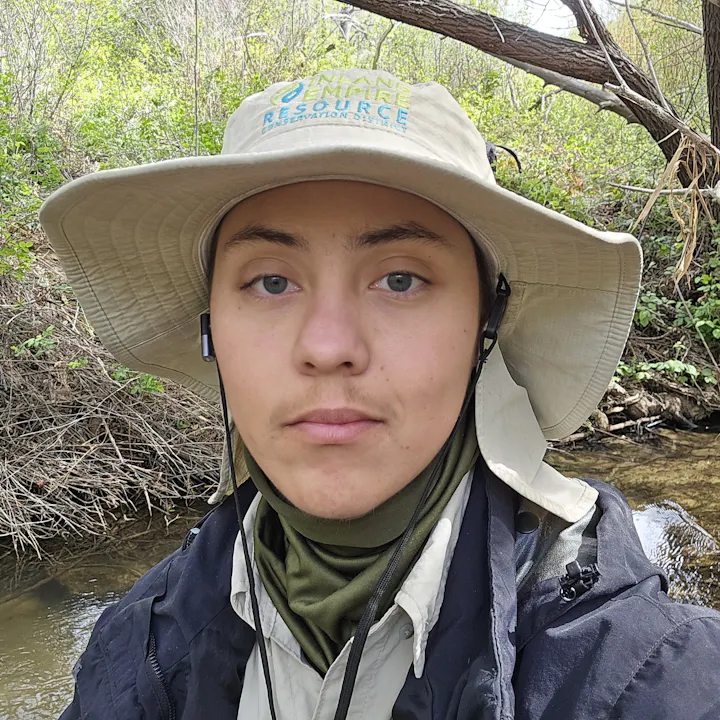 A person wearing a wide-brimmed hat and outdoor gear poses near a creek with greenery in the background.