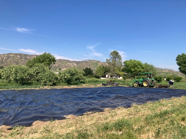 A tractor near a black tarp-covered field, surrounded by greenery and hills under a clear blue sky.