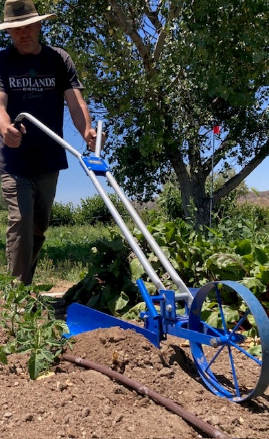 A person using a blue hand tiller in a garden, with greenery and trees in the background.