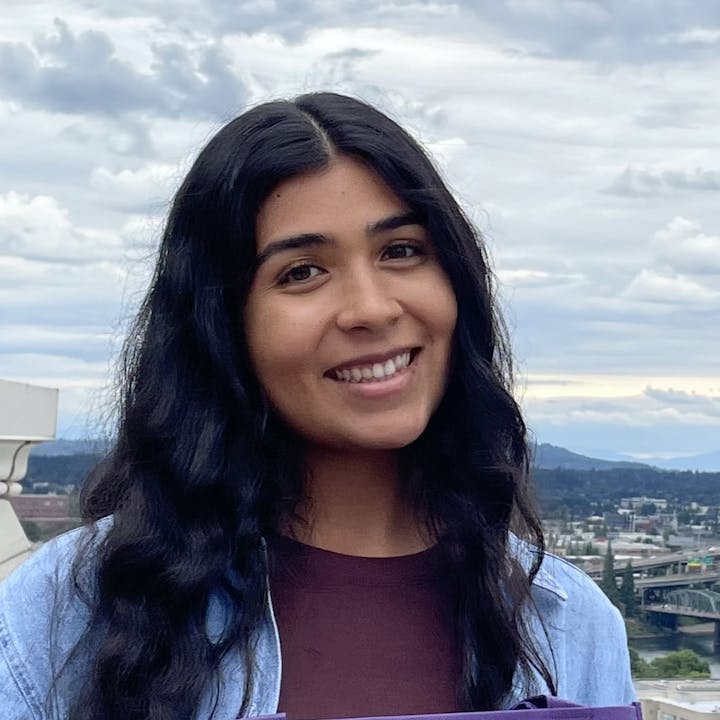 A smiling person with dark hair outdoors, cloudy sky and distance landscape in the background.
