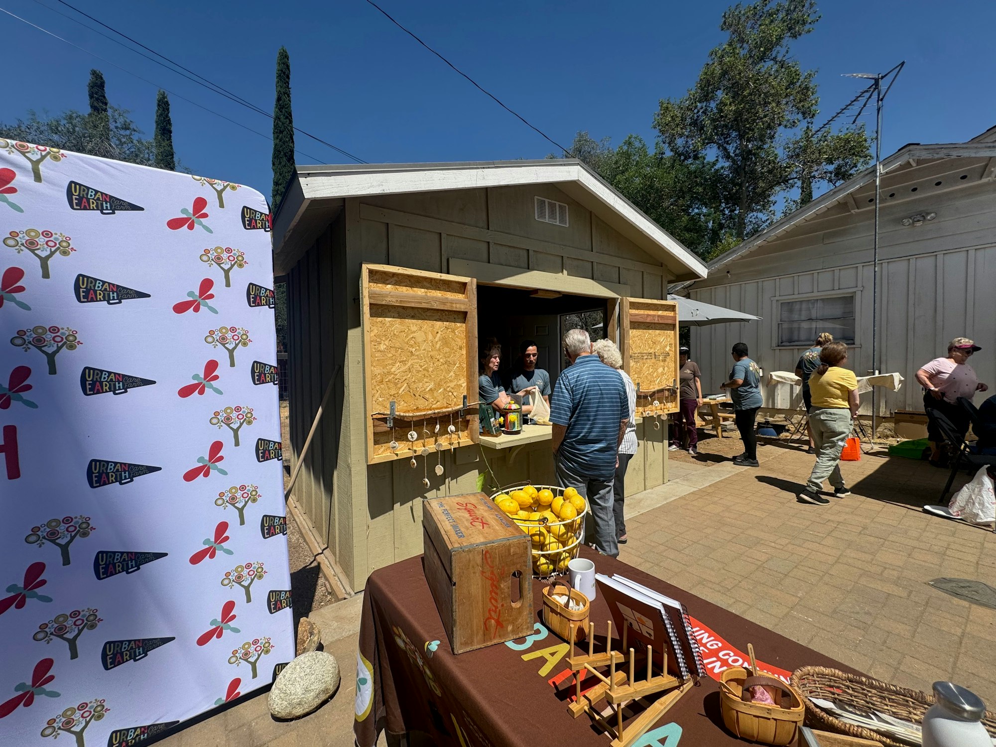 Outdoor market scene with a small building, people interacting, a banner, and a table with lemons and crafts.