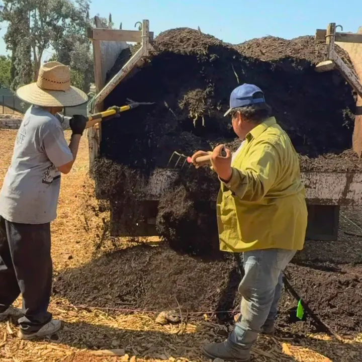Two people are working in a garden, using tools to handle compost or soil from a large truck. Sunny outdoor setting.