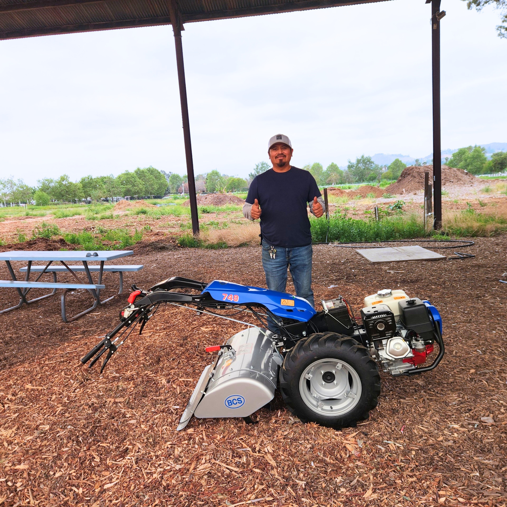 A person gives a thumbs-up next to a blue farming machine in a wooded area with scattered mulch and greenery in the background.