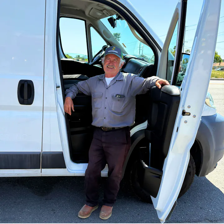 A man in work attire stands by the open door of a white van, smiling in a sunny outdoor setting.
