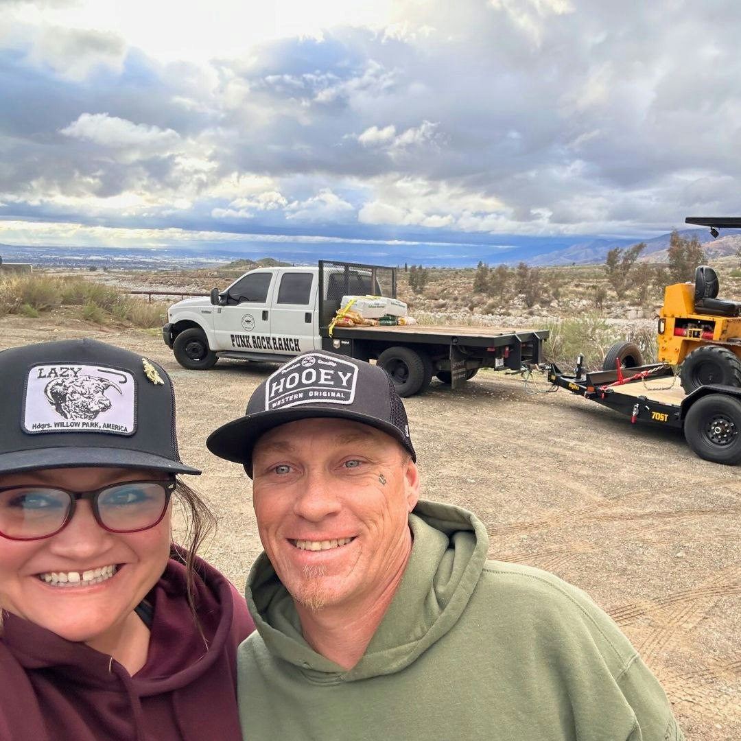 Two people smiling outdoors, a truck labeled "Funk Rock Ranch," trailer, and equipment in the background under cloudy skies.