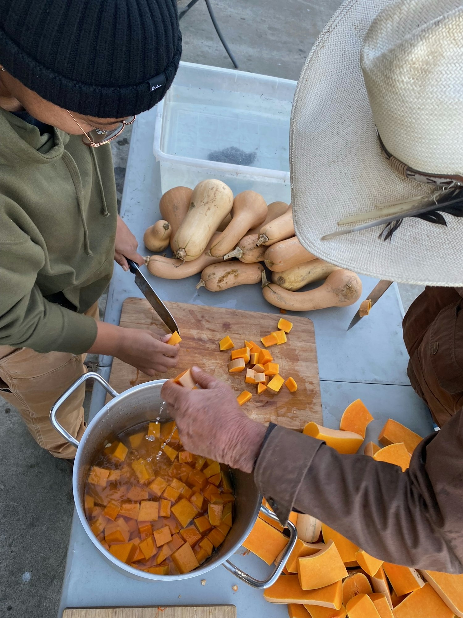 Two people are preparing butternut squash, chopping it for cooking, with a pot of diced squash and whole squash on the table.