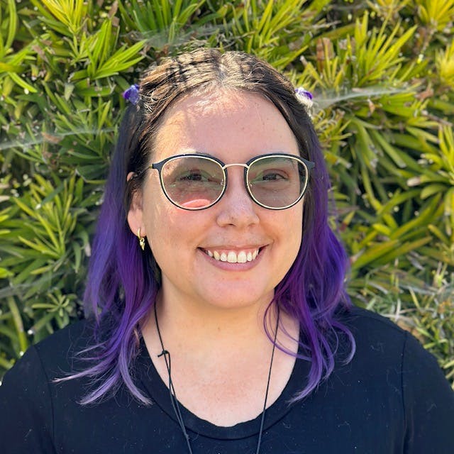 A smiling person with purple hair and glasses, standing in front of green plants.