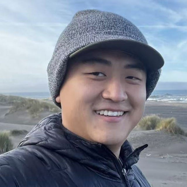 A smiling person in a beanie poses for a selfie at a beach, with waves and sand dunes in the background.
